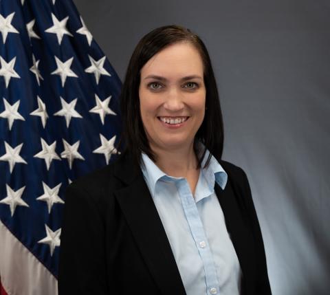 Image of a woman wearing a blue blouse with a black jacket looking at camera and smiling, standing in front of American flag.