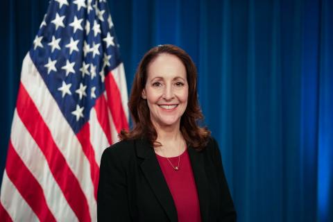 Photo of a woman in a black suit standing in front of a U.S. flag.